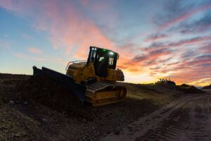 A Komatsu crawler dozer on a worksite at dusk, silhouetted against a dramatic sunset sky with orange and purple clouds.