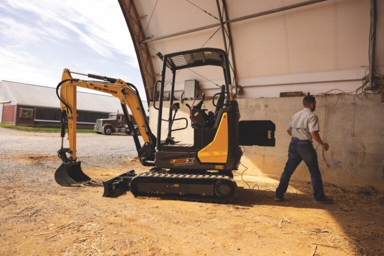 A New Holland Construction E25X electric mini excavator parked beside a farm building while an operator connects a charging cable to a wall-mounted socket