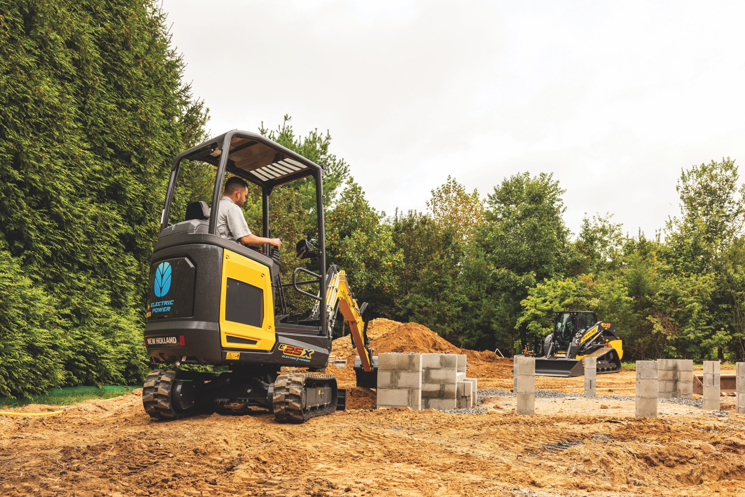 A New Holland Construction E25X electric mini excavator operated by a man in the foreground of an outdoor construction site, with a C314X electric mini track loader visible working in the background.