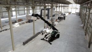 Alt text: Aerial view of a white and black Hidromek HMK 18-40 TH telehandler inside a large steel-framed industrial building under construction. The boom is fully extended and angled upward, with structural steelwork, equipment and building materials visible in the background.