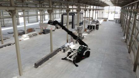 Alt text: Aerial view of a white and black Hidromek HMK 18-40 TH telehandler inside a large steel-framed industrial building under construction. The boom is fully extended and angled upward, with structural steelwork, equipment and building materials visible in the background.