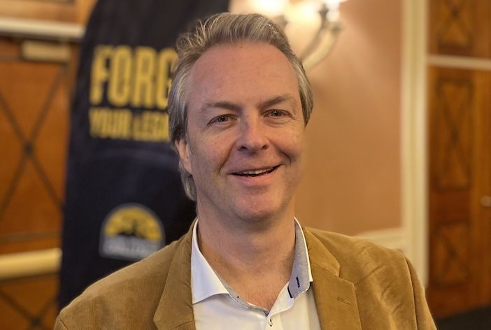 Herwig Peschl smiling at a conference event, wearing a tan blazer and open-collar white shirt, with a banner visible in the background.