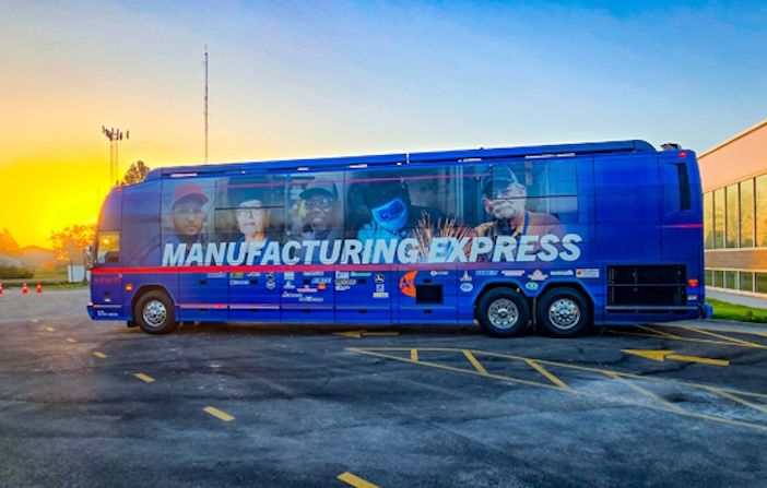A large blue touring coach wrapped in a full-vehicle graphic featuring the words "Manufacturing Express" in large white lettering, with images of manufacturing workers on the side panels and multiple sponsor logos along the lower edge, parked in a car park at sunset with a warm orange and yellow sky in the background.
