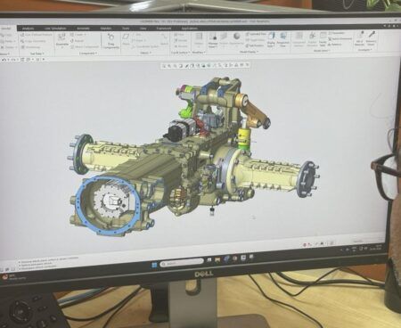An engineer wearing glasses views a detailed 3D CAD model of an axle and transmission assembly on a Dell monitor, displayed in Creo Parametric software. A Carraro Technologies India desk placard is visible in the foreground.
