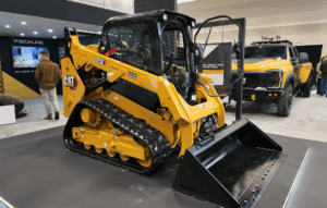 The Cat 253 compact track loader displayed on a stand at CONEXPO-CON/AGG, fitted with a dirt bucket and showing the full machine profile including cab, tracks and undercarriage. A VisionLink display and Cat compact track loader lineup board are visible in the background.