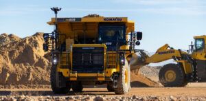 Front view of a yellow Komatsu HD605 haul truck fitted with autonomous system sensors and computing hardware mounted on the cab and front structure, at a dusty open-pit quarry site. A Komatsu wheel loader is visible to the right in the background.
