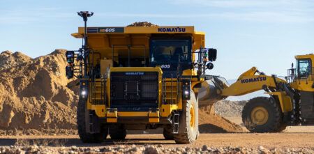 Front view of a yellow Komatsu HD605 haul truck fitted with autonomous system sensors and computing hardware mounted on the cab and front structure, at a dusty open-pit quarry site. A Komatsu wheel loader is visible to the right in the background.