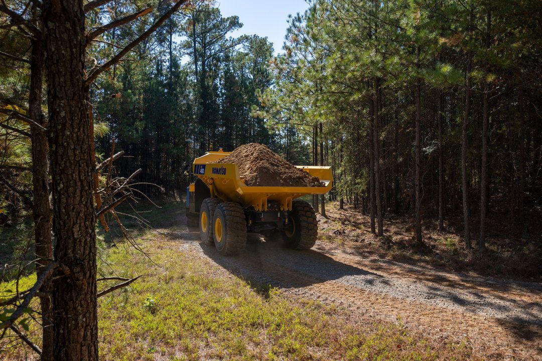 A yellow Komatsu HM460-6 articulated dump truck loaded with soil travelling along a narrow dirt track through a pine forest.
