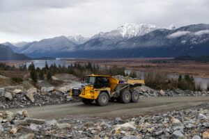 A yellow Komatsu HM460-6 articulated haul truck travelling along a rocky haul road with snow-capped mountains and a river valley visible in the background