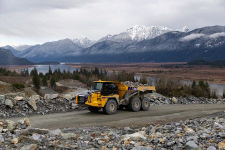 A yellow Komatsu HM460-6 articulated haul truck travelling along a rocky haul road with snow-capped mountains and a river valley visible in the background