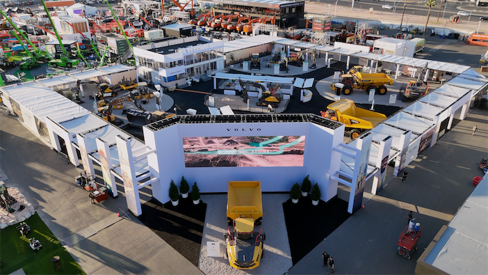 Aerial drone view of the Volvo Construction Equipment outdoor exhibition booth at ConExpo-Con/Agg 2026 in Las Vegas, showing a large white-canopied stand with multiple yellow construction machines on display, including articulated haulers and wheel loaders, with a large Volvo-branded LED screen visible at the entrance.