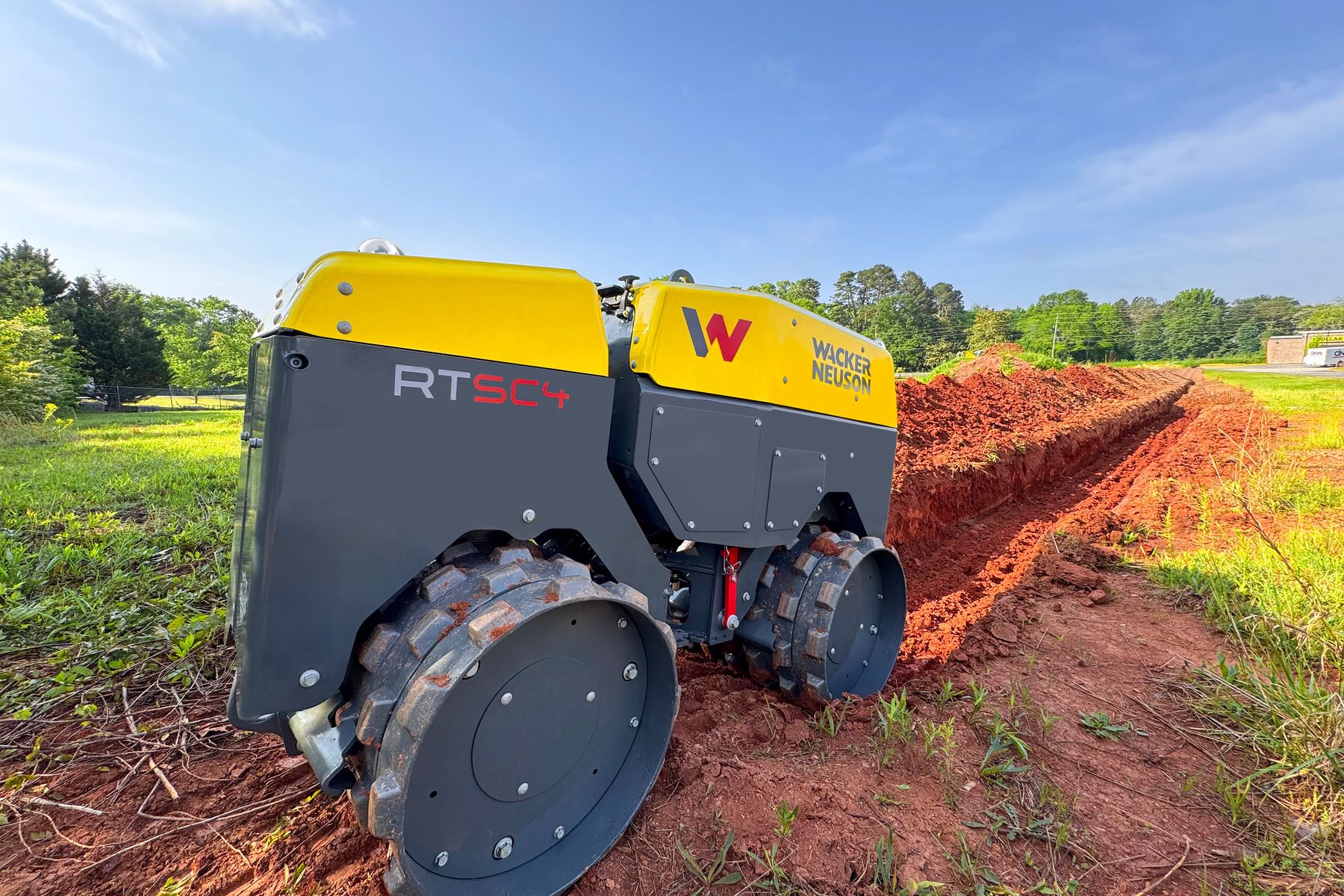 A yellow and grey Wacker Neuson RTsc4 remote-controlled trench roller working in a red clay trench on a construction site