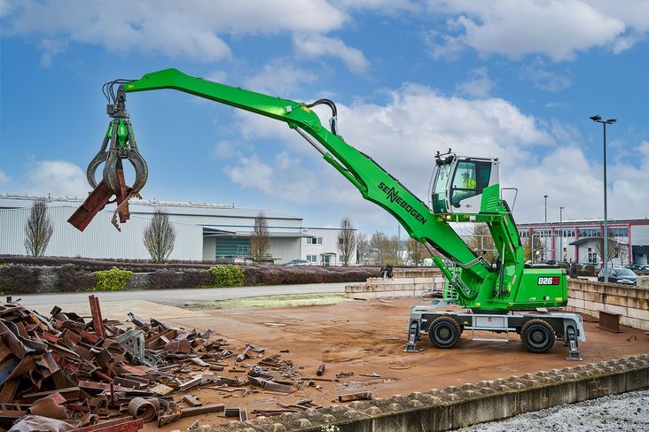 A green Sennebogen 826 G Electro Battery wheeled material handler positioned on a concrete pad at an outdoor recycling facility. The machine's orange peel grapple attachment is raised and partially open, holding scrap metal, with a pile of mixed scrap visible to the left and industrial buildings in the background.