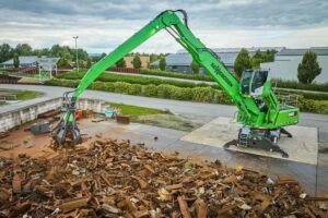 A green Sennebogen 830 G wheeled material handler positioned on a concrete platform above a large pile of mixed scrap metal at an outdoor recycling facility. The machine's grapple attachment is extended at the end of the compact boom, with industrial buildings, hedgerows and trees visible in the background under an overcast sky.