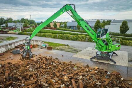 Sennebogen to showcase recycling and demolition range at IFAT 2026 A green Sennebogen 830 G wheeled material handler positioned on a concrete platform above a large pile of mixed scrap metal at an outdoor recycling facility. The machine's grapple attachment is extended at the end of the compact boom, with industrial buildings, hedgerows and trees visible in the background under an overcast sky.