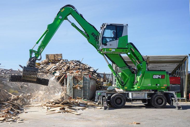 A green Sennebogen 824 G Electro Battery wheeled material handler working at a recycling site, using a grapple attachment to move a load of mixed timber and wood waste. The elevated cab is visible above the machine's upper structure, with a corrugated metal building and additional material stockpiles in the background under a clear blue sky.