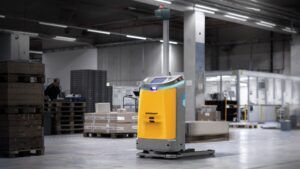 A Jungheinrich EAE 212a autonomous mobile robot with a yellow and grey body, blue indicator light and tall sensor mast, operating in a warehouse environment. Wooden pallets, cardboard boxes and a worker handling goods are visible in the background.