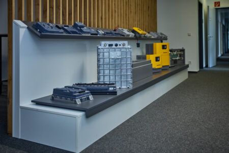 A display bench showing a range of Jungheinrich battery modules, chargers and power electronics components arranged on two tiers against a wood-panelled wall