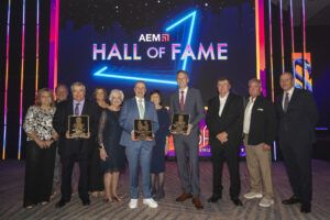 A group of people standing on stage at the AEM Hall of Fame ceremony, holding commemorative plaques, with the AEM Hall of Fame logo illuminated on a screen behind them