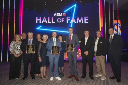A group of people standing on stage at the AEM Hall of Fame ceremony, holding commemorative plaques, with the AEM Hall of Fame logo illuminated on a screen behind them