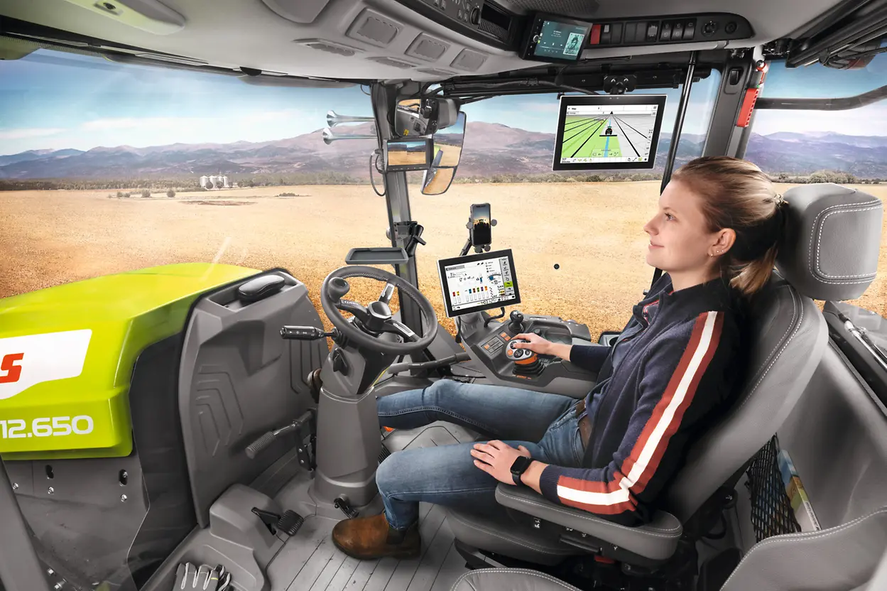  Female operator seated in the Claas Xerion 12 cab using the armrest-mounted control terminal, with a guidance display in front and a field visible through the windscreen
