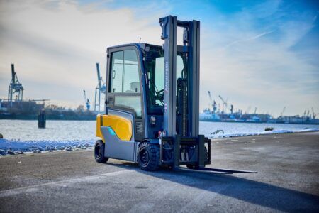 A Jungheinrich electric counterbalance forklift truck in yellow and grey livery, shown from the front-left angle on a concrete quayside. The truck has its forks lowered and the mast is visible in the foreground. In the background, Hamburg harbour is visible with port cranes, ships and a snow-dusted shoreline under a partly cloudy sky