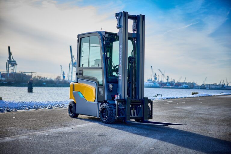 A Jungheinrich electric counterbalance forklift truck in yellow and grey livery, shown from the front-left angle on a concrete quayside. The truck has its forks lowered and the mast is visible in the foreground. In the background, Hamburg harbour is visible with port cranes, ships and a snow-dusted shoreline under a partly cloudy sky