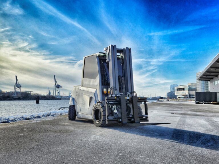 A Jungheinrich FalcOn electric forklift truck photographed on an industrial waterfront site. The vehicle has an angular, modern silver-grey body design with large industrial tyres and a heavy-duty mast and fork assembly. Port cranes and modern buildings are visible in the background under a blue sky with wispy clouds