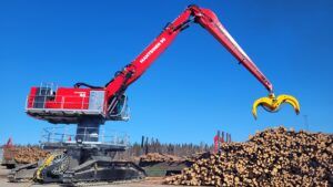 A red Mantsinen 90 material handler on crawler tracks operating at a log yard beside a large red industrial building. The machine's boom is fully extended, reaching over a tall pile of stacked logs with a yellow grapple visible at the tip. A second smaller material handler is visible in the background, and the site is set against a clear blue sky with coniferous forest on the horizon.