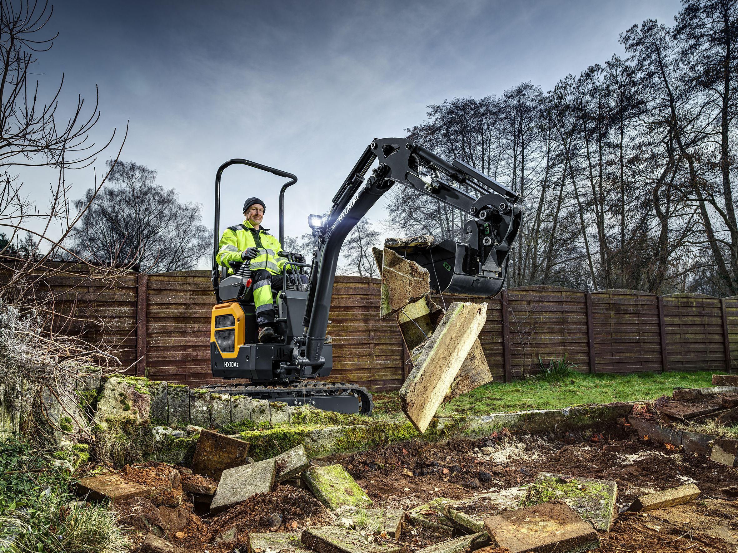Hyundai HX10A Z micro excavator with operator lifting a broken paving slab from a garden demolition site, with timber fencing in the background
