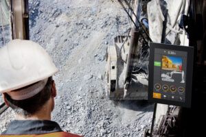 View from inside an excavator cab looking down into a rocky quarry pit, with an operator wearing a white hard hat and high-visibility vest in the foreground. A CrossControl CCpilot display is mounted to the right of the cab, showing a camera feed of a haul truck, a machine schematic, diagnostic gauges for RPM, engine temperature, fuel level and voltage, and navigation icons at the bottom of the screen.