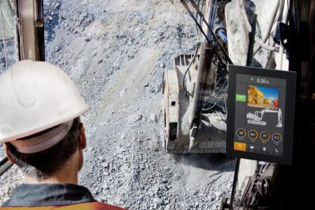 CrossControl introduces display computers with integrated Ethernet switch View from inside an excavator cab looking down into a rocky quarry pit, with an operator wearing a white hard hat and high-visibility vest in the foreground. A CrossControl CCpilot display is mounted to the right of the cab, showing a camera feed of a haul truck, a machine schematic, diagnostic gauges for RPM, engine temperature, fuel level and voltage, and navigation icons at the bottom of the screen.
