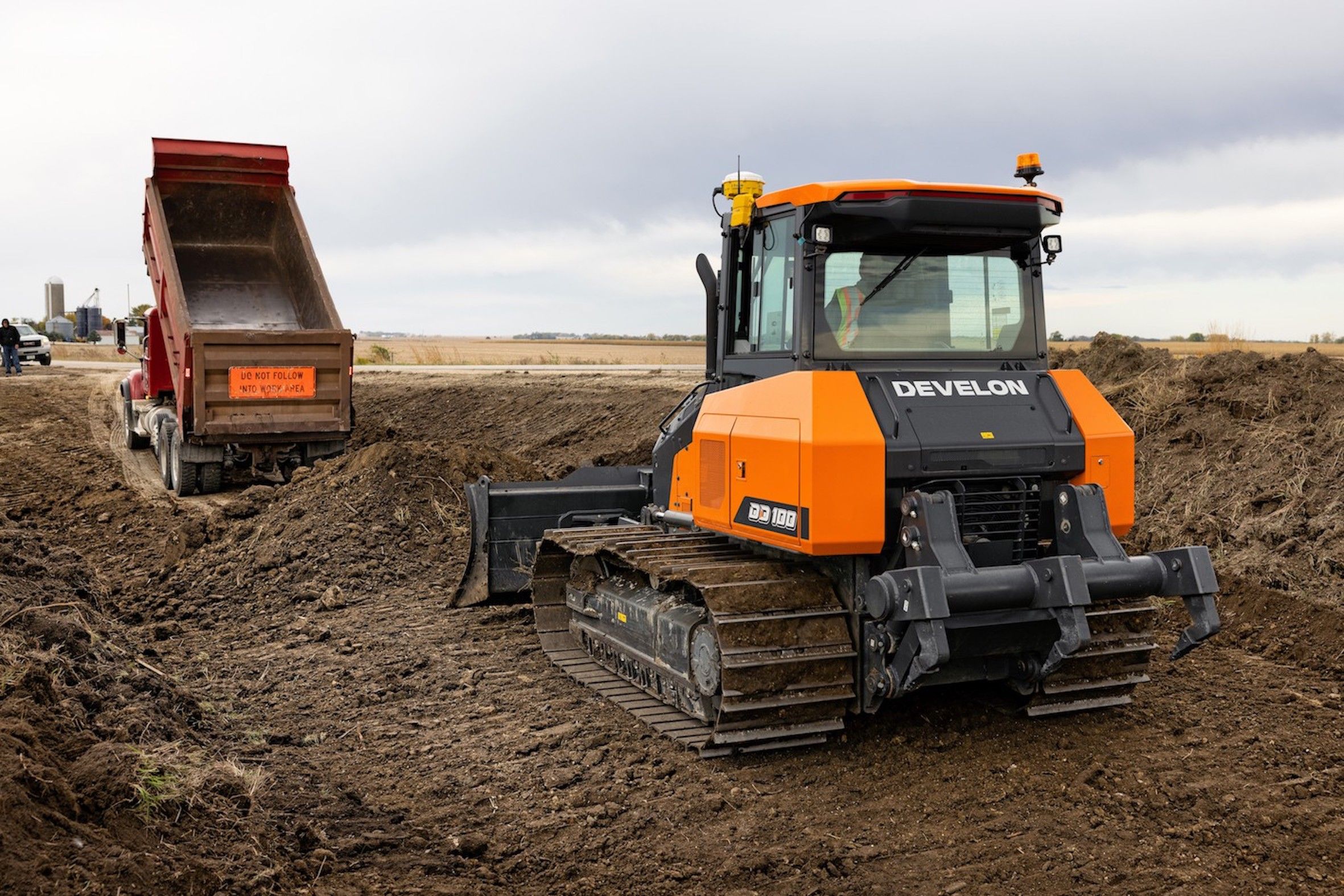 A rear-right view of an orange and grey Develon DD100 dozer operating on a muddy construction site, with a red dump truck with its bed raised visible in the background. An operator wearing a high-visibility vest is seated in the cab. The flat rural landscape and overcast sky are visible beyond the site.