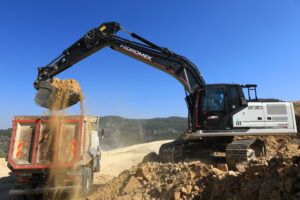 A black and white Hidromek HMK 230 LC crawler excavator tipping a bucket of earth into a dump truck at a quarry, with hills in the background