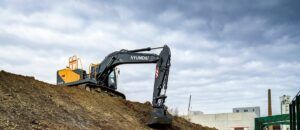 A Hyundai HX230 crawler excavator in yellow and dark grey livery, positioned on a mound of earth at a construction site with an overcast sky in the background.