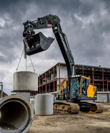 A Hyundai HX230 crawler excavator using chains to lift a large concrete pipe section, with additional concrete pipes and a steel-framed building under construction visible in the background