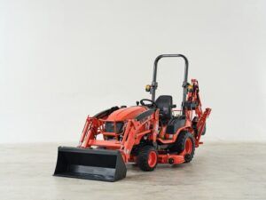 A Kioti sub-compact tractor in the manufacturer's signature orange livery, photographed in a studio setting against a neutral background. The tractor is fitted with a front-end loader bucket and a rear backhoe attachment, and features a ROPS (roll-over protective structure) with a high-back operator seat. The compact machine sits on turf-style tyres.