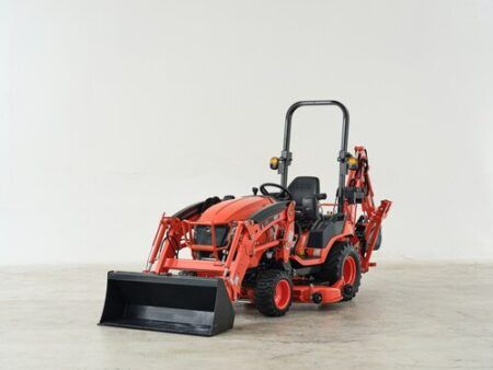 A Kioti sub-compact tractor in the manufacturer's signature orange livery, photographed in a studio setting against a neutral background. The tractor is fitted with a front-end loader bucket and a rear backhoe attachment, and features a ROPS (roll-over protective structure) with a high-back operator seat. The compact machine sits on turf-style tyres.