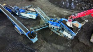 Aerial photograph of a Kleemann mobile crushing and screening plant train on a jobsite, showing two blue Kleemann machines – a scalper and an impact crusher – with discharge conveyors, being fed by a red excavator. Processed material is discharged onto a stockpile via a mobile belt conveyor