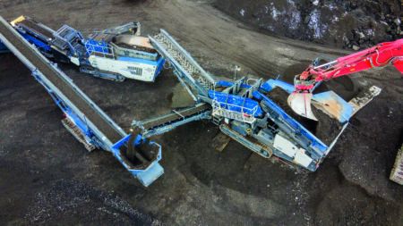 Aerial photograph of a Kleemann mobile crushing and screening plant train on a jobsite, showing two blue Kleemann machines – a scalper and an impact crusher – with discharge conveyors, being fed by a red excavator. Processed material is discharged onto a stockpile via a mobile belt conveyor