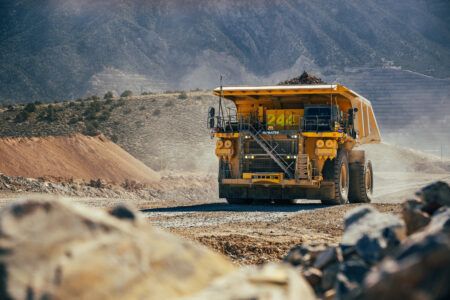 A large yellow Komatsu 930E-5AT autonomous haul truck loaded with ore travels along a haul road at Barrick's Nevada Gold Mines open-pit operation, with rocky mountain terrain in the background