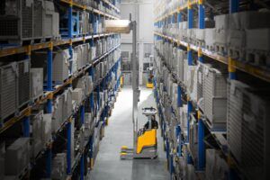An operator driving a yellow Jungheinrich ETV 4i reach truck between tall pallet racking in a warehouse, with the mast raised to retrieve a pallet from an upper level