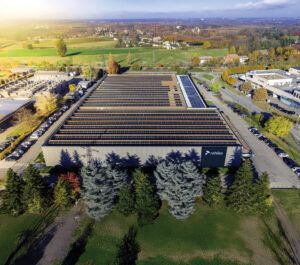 An aerial photograph of Rehlko's Reggio Emilia facility, showing a large industrial building with extensive rooftop solar panel arrays. The Rehlko logo is visible on the front facade of the building. The facility is surrounded by car parks, mature trees and green countryside, with residential areas visible in the background under hazy sunlight.