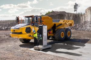 A yellow Volvo A40 Electric articulated hauler connected to a charging unit at a quarry, with an operator in high-visibility clothing handling the charging cable