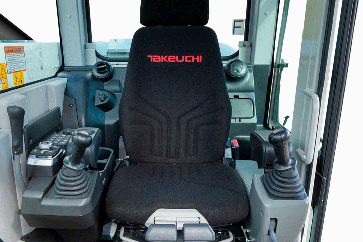  Interior view of the Takeuchi TB3150 series excavator cab showing a black air suspension operator's seat with the Takeuchi logo, flanked by pilot-operated joystick controls and a multi-switch control panel on the left console