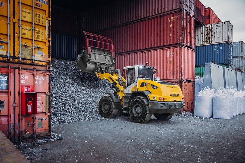 A yellow Liebherr L 546 wheel loader tipping a bucket of crushed material between shipping containers at a recycling yard