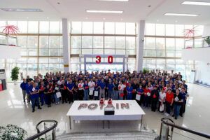 A large group of employees in blue and red shirts assembled in a factory entrance hall, with a "30" balloon display and Potain-branded table in the foreground