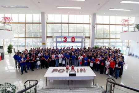 A large group of employees in blue and red shirts assembled in a factory entrance hall, with a "30" balloon display and Potain-branded table in the foreground