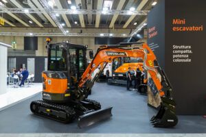 Photograph of an orange and black Develon DX27Z-7K mini excavator displayed at an indoor exhibition stand, with a larger crawler excavator visible in the background beneath signage reading "Mini escavatori".