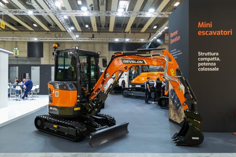 Photograph of an orange and black Develon DX27Z-7K mini excavator displayed at an indoor exhibition stand, with a larger crawler excavator visible in the background beneath signage reading "Mini escavatori".